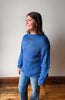 A woman wearing a blue chunky knit sweater and blue jeans, standing in a room with a light gray wall and wooden floor.