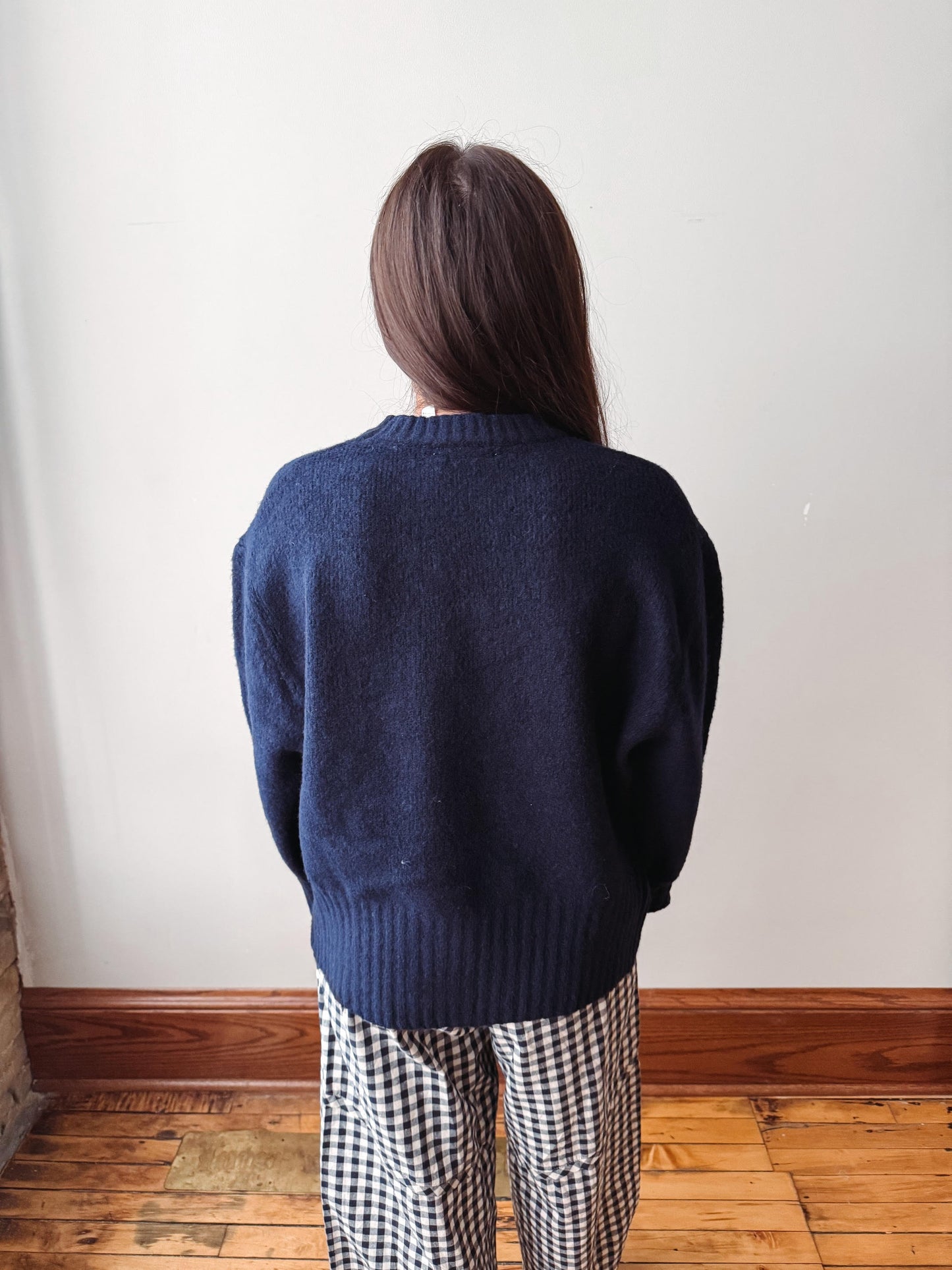 A person viewed from behind wearing a dark navy blue knit sweater and black-and-white gingham pants, standing against an off-white wall with a wooden baseboard and wood floor.