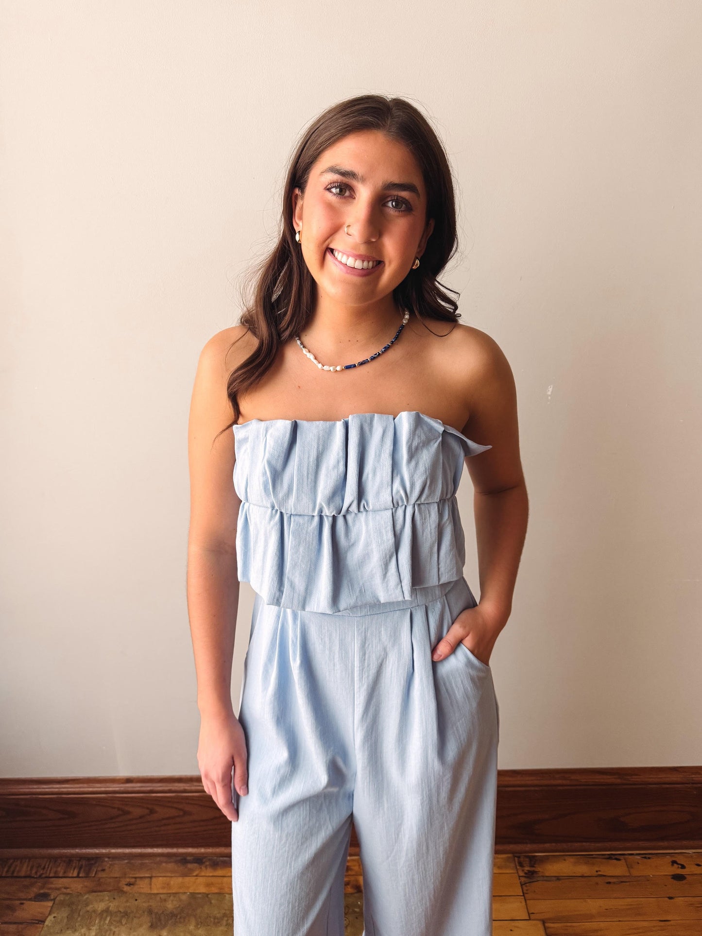 Woman wearing a light blue strapless jumpsuit standing against a plain wall.
