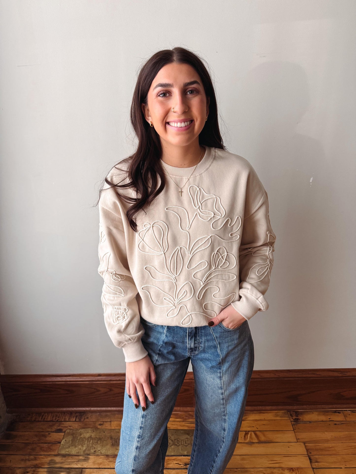 A woman with long dark hair stands indoors wearing a beige crewneck sweatshirt with white floral embroidery and blue jeans, posing against a plain off-white wall with a wooden floor visible.