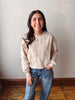 A woman with long dark hair stands indoors wearing a beige crewneck sweatshirt with white floral embroidery and blue jeans, posing against a plain off-white wall with a wooden floor visible.