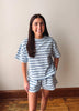 A woman modeling a matching two-piece loungewear set consisting of a short-sleeve t-shirt and shorts, both featuring a light blue and white horizontal stripe pattern, set against an indoor wall background.