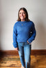 A woman wearing a blue ribbed knit sweater and blue jeans, standing in a room with a light gray wall and wooden floor.