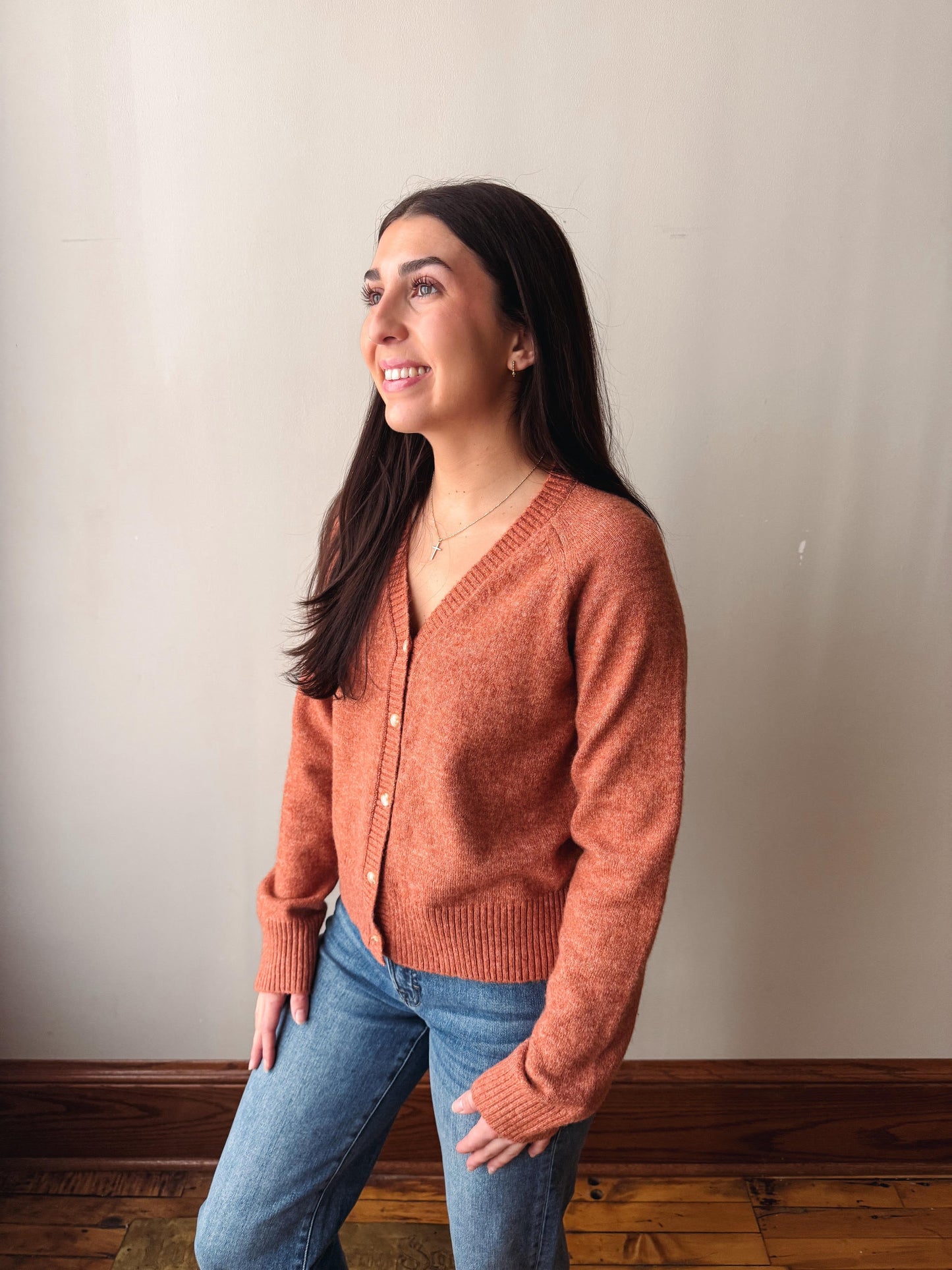 A woman wearing a rust-orange knit cardigan sweater and blue jeans, standing against a plain beige wall with a wooden floor visible.