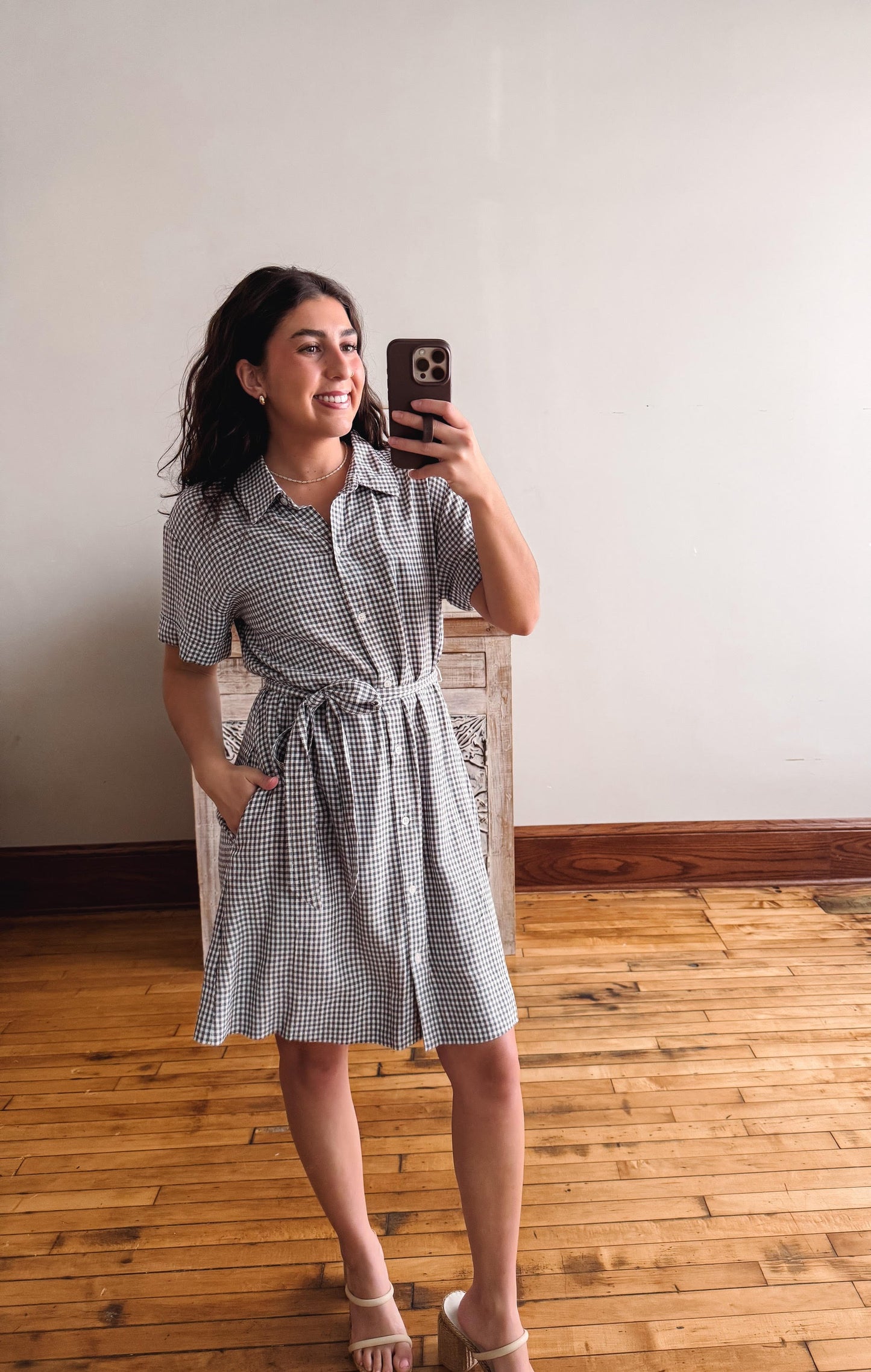Woman taking a mirror selfie wearing a checkered dress in a room with wooden flooring.