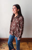 A woman wearing a brown long-sleeve button-up shirt with a white horse pattern, paired with blue jeans, standing in a room with a light gray wall and wooden floor.