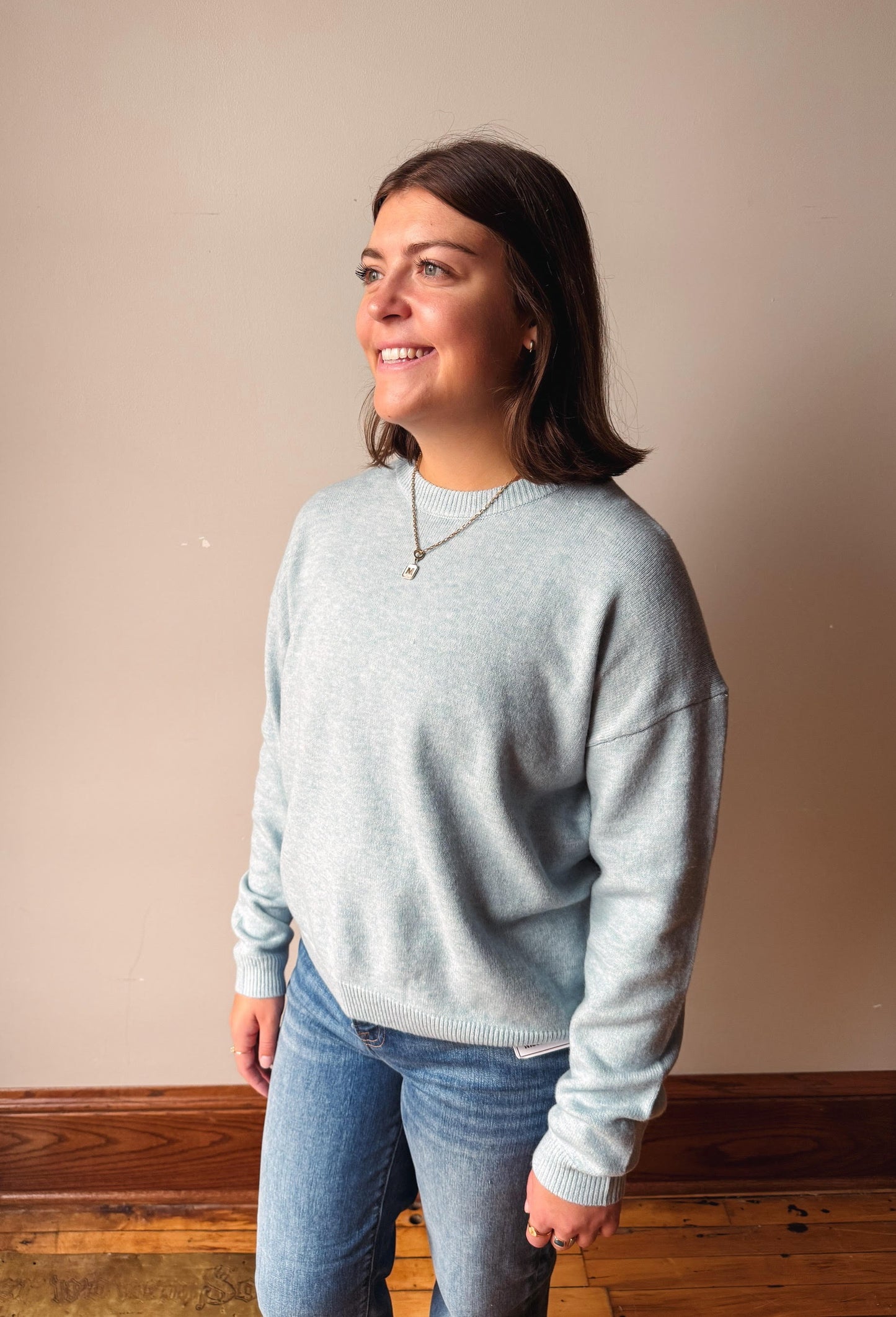 A woman wearing a light heather gray crewneck sweatshirt and blue jeans, standing against a plain beige wall with wooden trim and a wood floor.