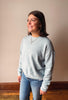 A woman wearing a light heather gray crewneck sweatshirt and blue jeans, standing against a plain beige wall with wooden trim and a wood floor.