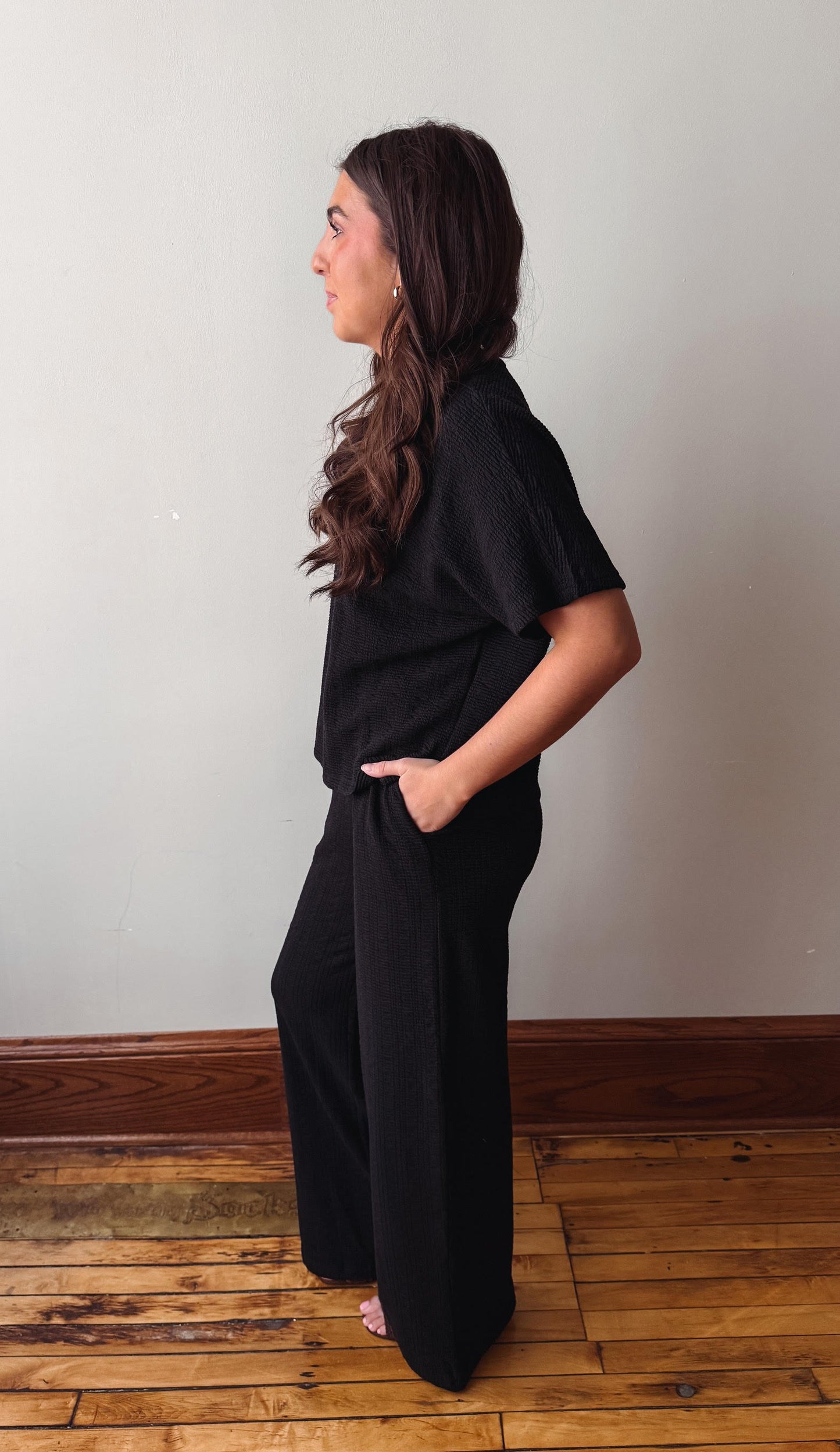 Woman wearing a black outfit standing on a wooden floor against a white wall.