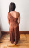 A woman viewed from behind wearing a brown one-shoulder dress with white polka dots, standing on a wooden floor against a plain white wall.
