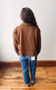 A person viewed from behind wearing a brown suede jacket, blue jeans, and brown boots, standing on a wooden floor against a plain off-white wall.