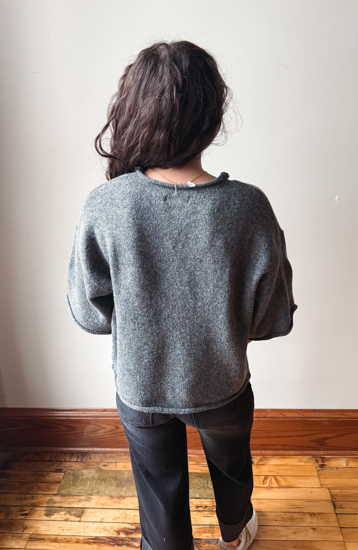 A person viewed from behind wearing a gray knit sweater and dark pants, standing on a wooden floor against a plain off-white wall.