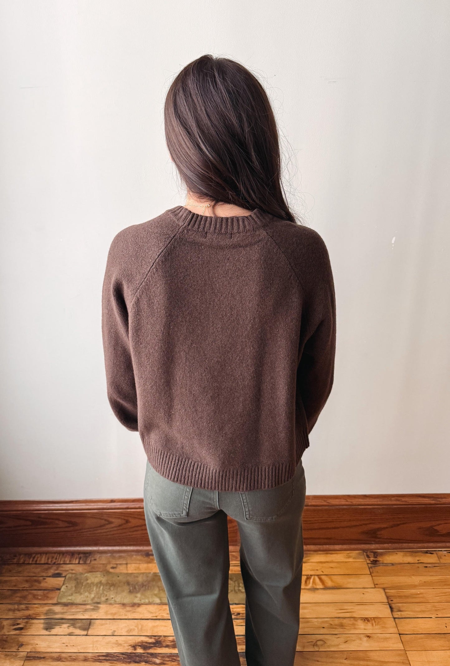 A woman viewed from behind wearing a brown knit crewneck sweater and olive green pants, standing on a wooden floor against a plain off-white wall.