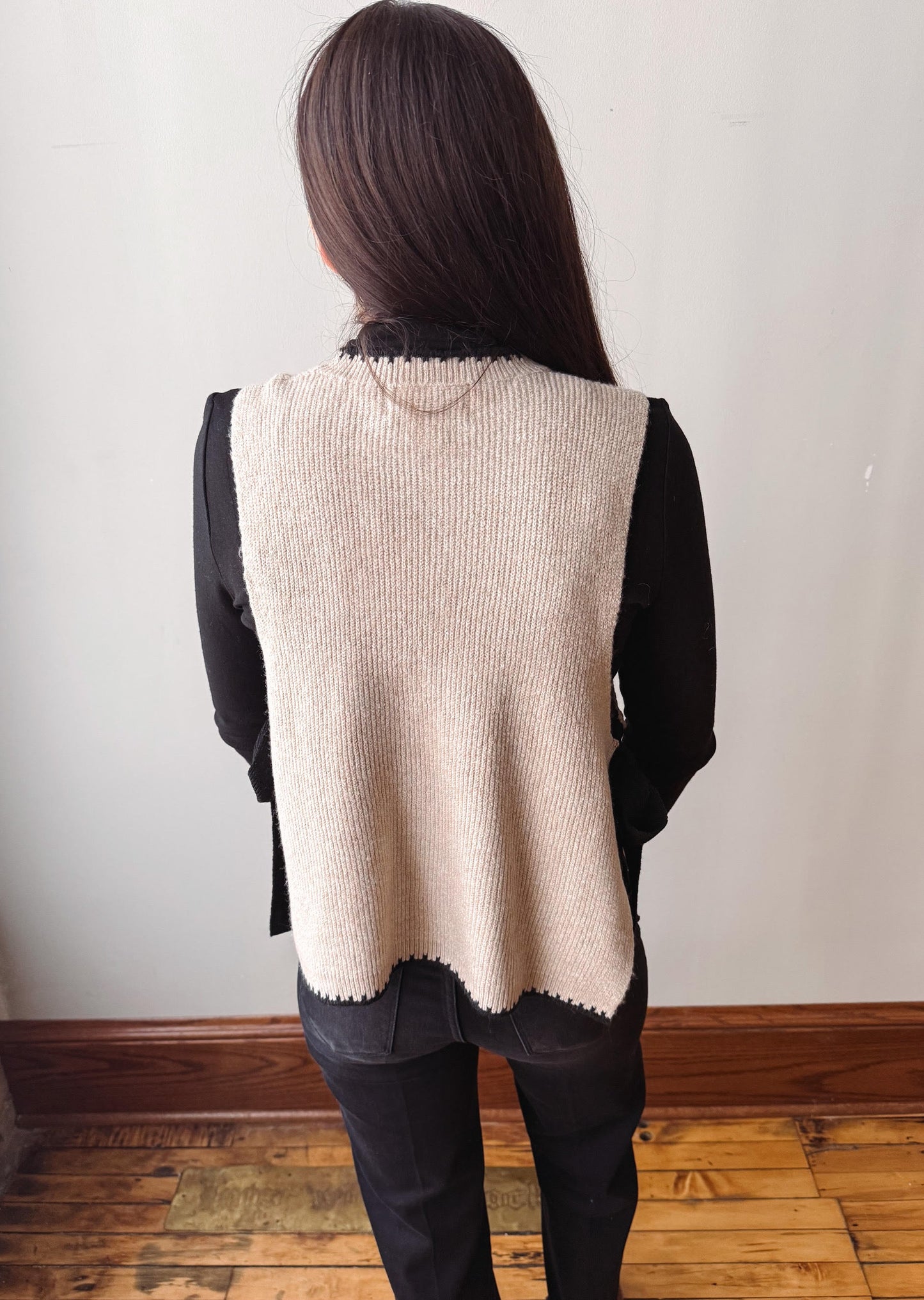 A woman viewed from behind wearing a beige ribbed knit sleeveless vest over a black long-sleeve top and black pants, standing on a wooden floor against a plain light wall.