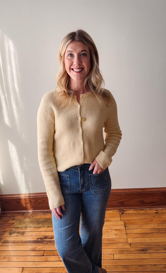 Woman wearing a yellow cardigan and blue jeans standing in a room with wooden flooring and a white wall.