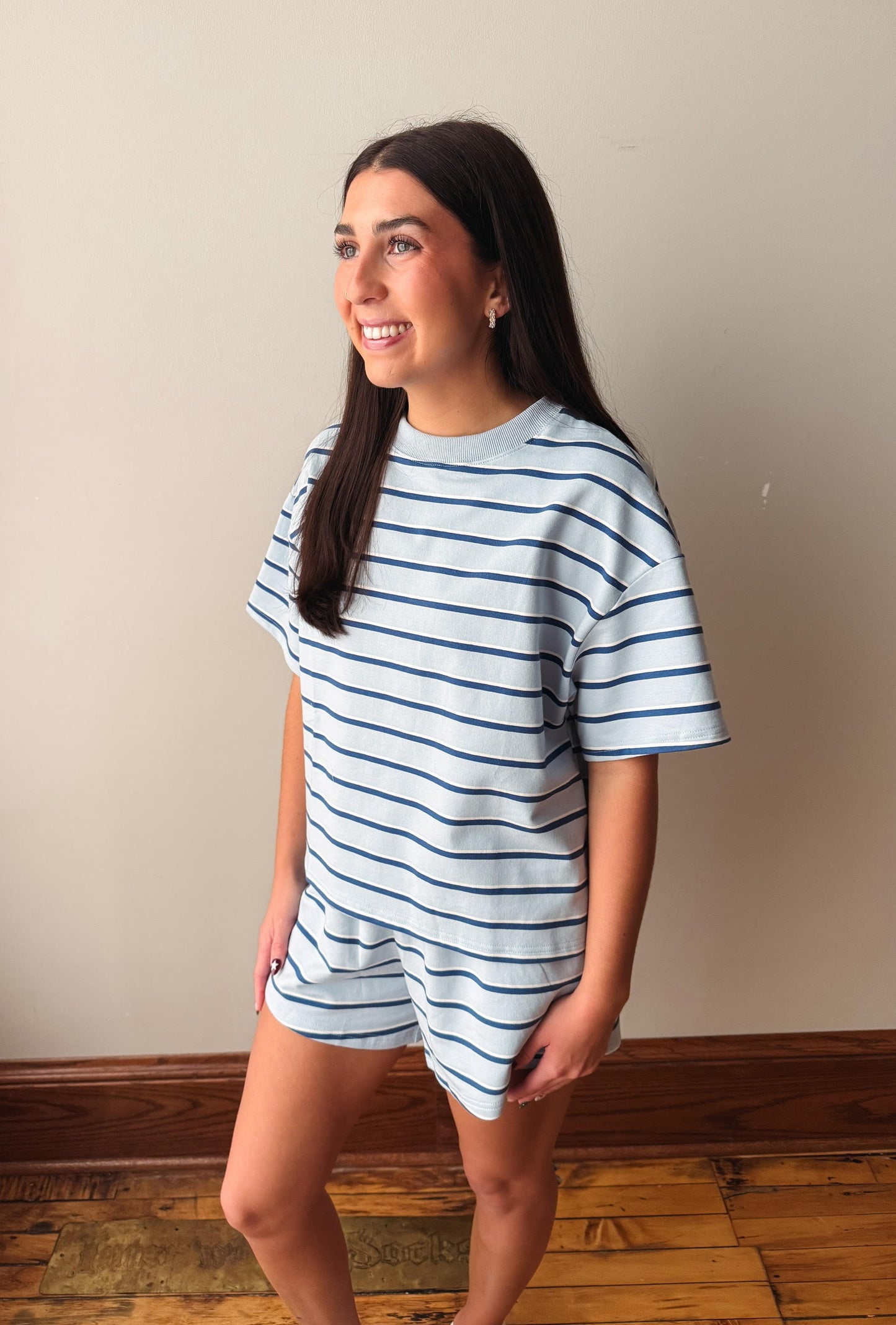 A woman modeling a light blue and navy blue horizontally striped short-sleeve t-shirt and matching shorts set, standing against a beige wall with wooden flooring.