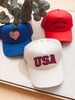 Three baseball caps with 'USA' and 'American Crown' text on a light surface with greenery.