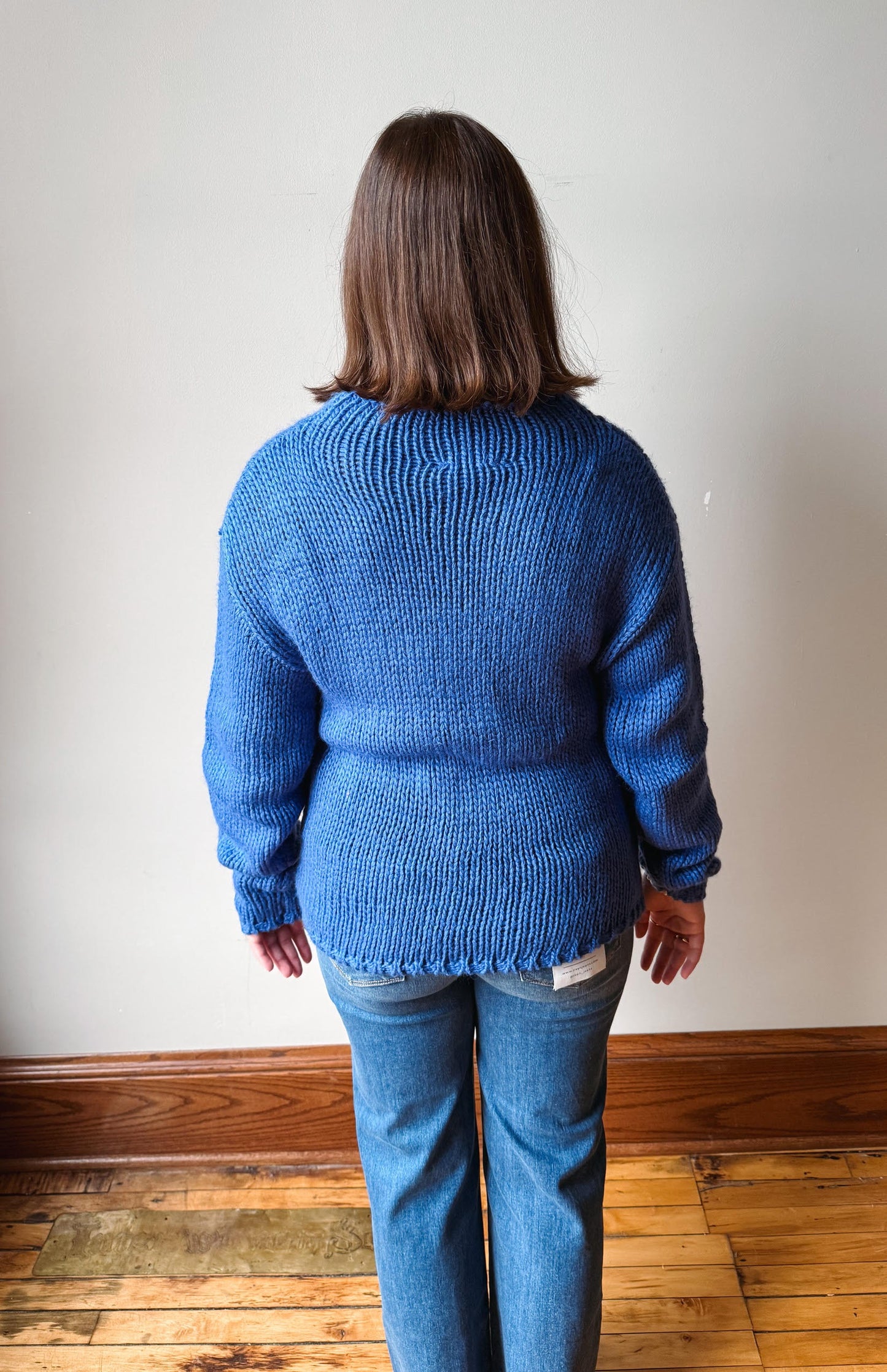 A woman viewed from behind wearing a blue ribbed knit sweater and blue jeans, standing on a wooden floor against a plain off-white wall.