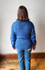 A woman viewed from behind wearing a blue ribbed knit sweater and blue jeans, standing on a wooden floor against a plain off-white wall.