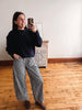 A woman takes a mirror selfie in a room with hardwood floors and a white wall, wearing a dark navy long-sleeve sweater and black-and-white gingham wide-leg pants.