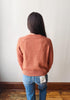 A person viewed from behind wearing a textured, heathered terracotta orange knit sweater and blue jeans, standing against a plain off-white wall with a wooden baseboard and floor.