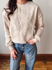 A person wearing a cream-colored crewneck sweatshirt with large floral embroidery on the front, paired with blue jeans, standing against a neutral wall.
