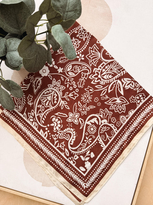 A square bandana with a white paisley and floral print on a rust red background, styled with green eucalyptus leaves on a light beige surface.