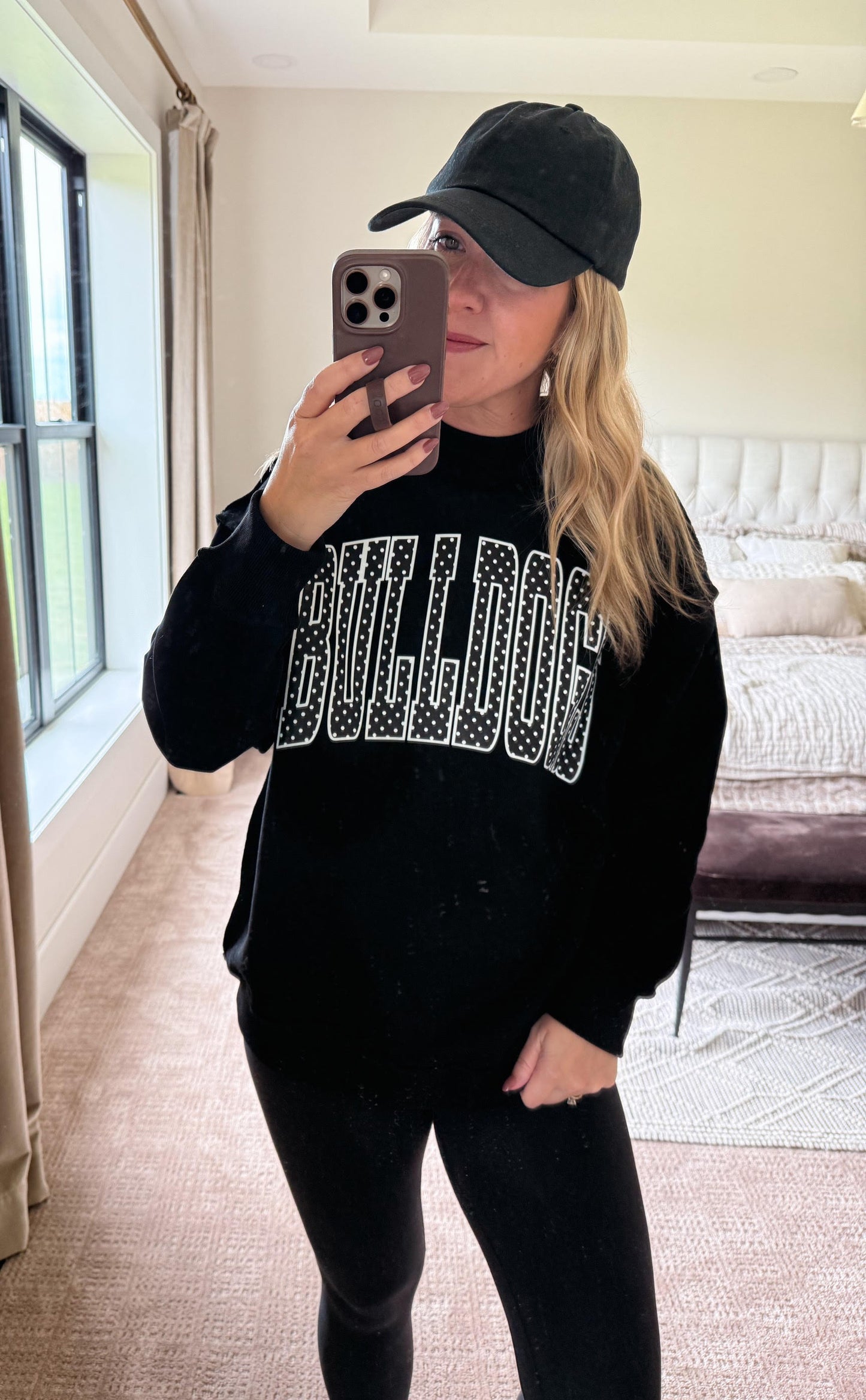 A woman takes a mirror selfie in a bedroom while wearing a black crewneck sweatshirt with 'BULLDOG' printed across the chest in sparkling silver lettering, paired with a black baseball cap and black leggings.