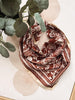 A rust brown and cream paisley bandana scarf styled with green eucalyptus leaves on a beige background with circular patterns.
