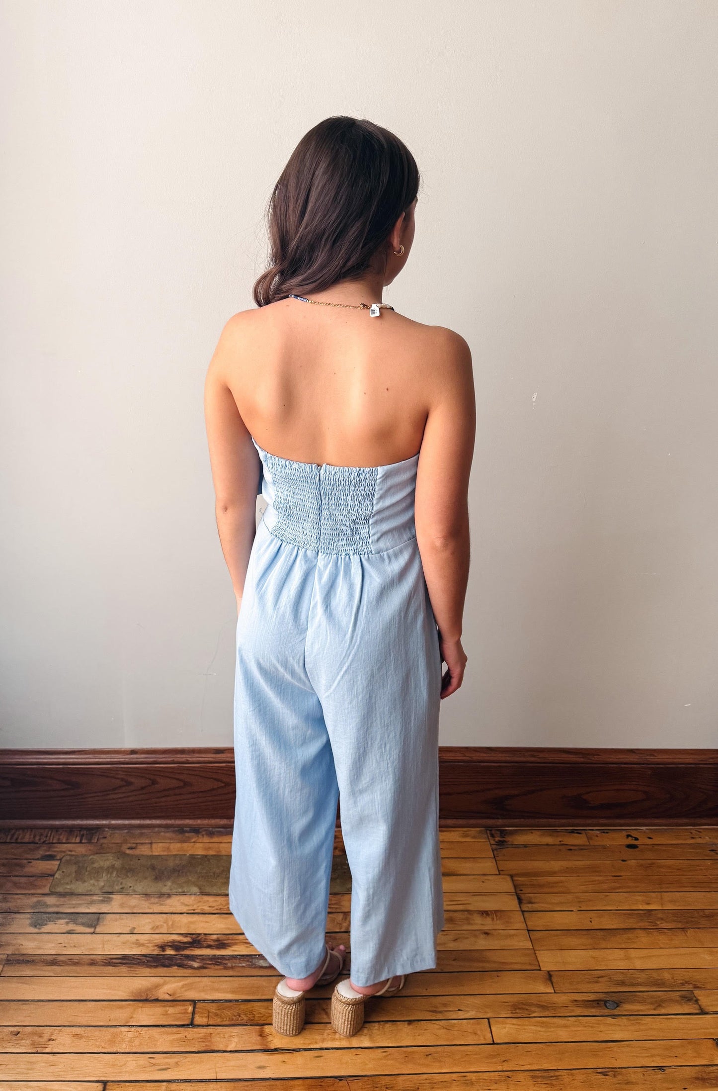 Woman wearing a light blue strapless jumpsuit standing on a wooden floor against a plain wall.