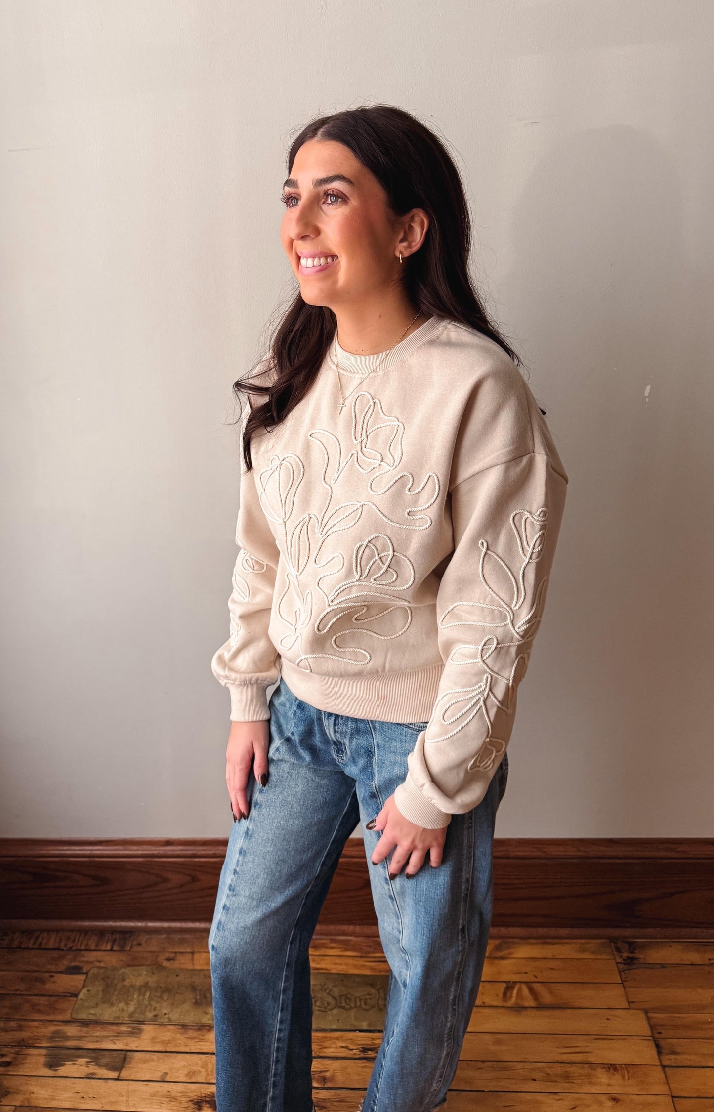 A woman wearing a cream-colored crewneck sweatshirt with intricate floral embroidery on the front and sleeves, paired with blue jeans, standing in a room with a light gray wall and wooden floor.