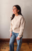 A woman wearing a cream-colored crewneck sweatshirt with intricate floral embroidery on the front and sleeves, paired with blue jeans, standing in a room with a light gray wall and wooden floor.