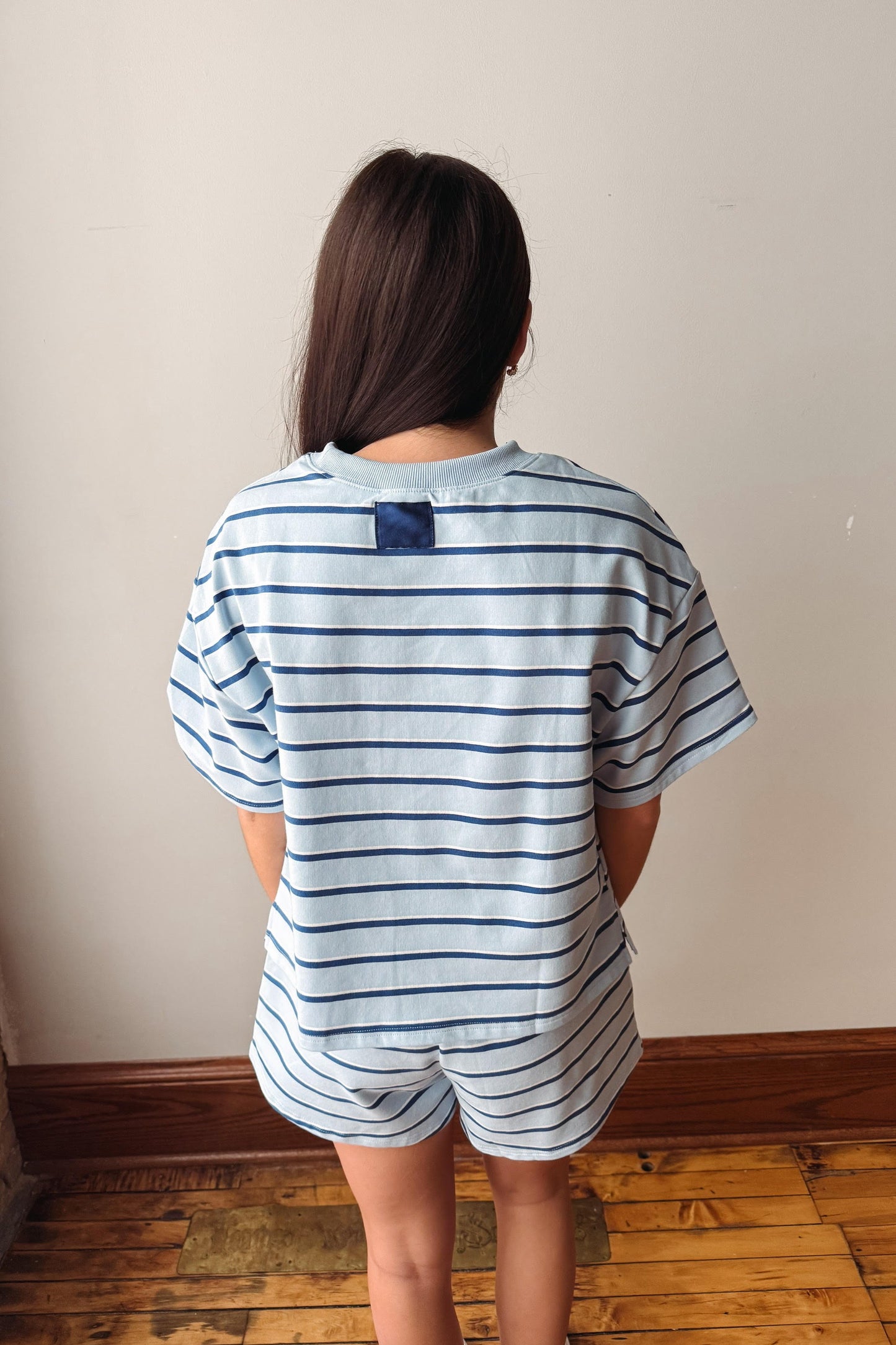 A person viewed from behind wearing a light blue and navy blue horizontally striped short-sleeve t-shirt and matching shorts, standing on a wooden floor against a plain beige wall.