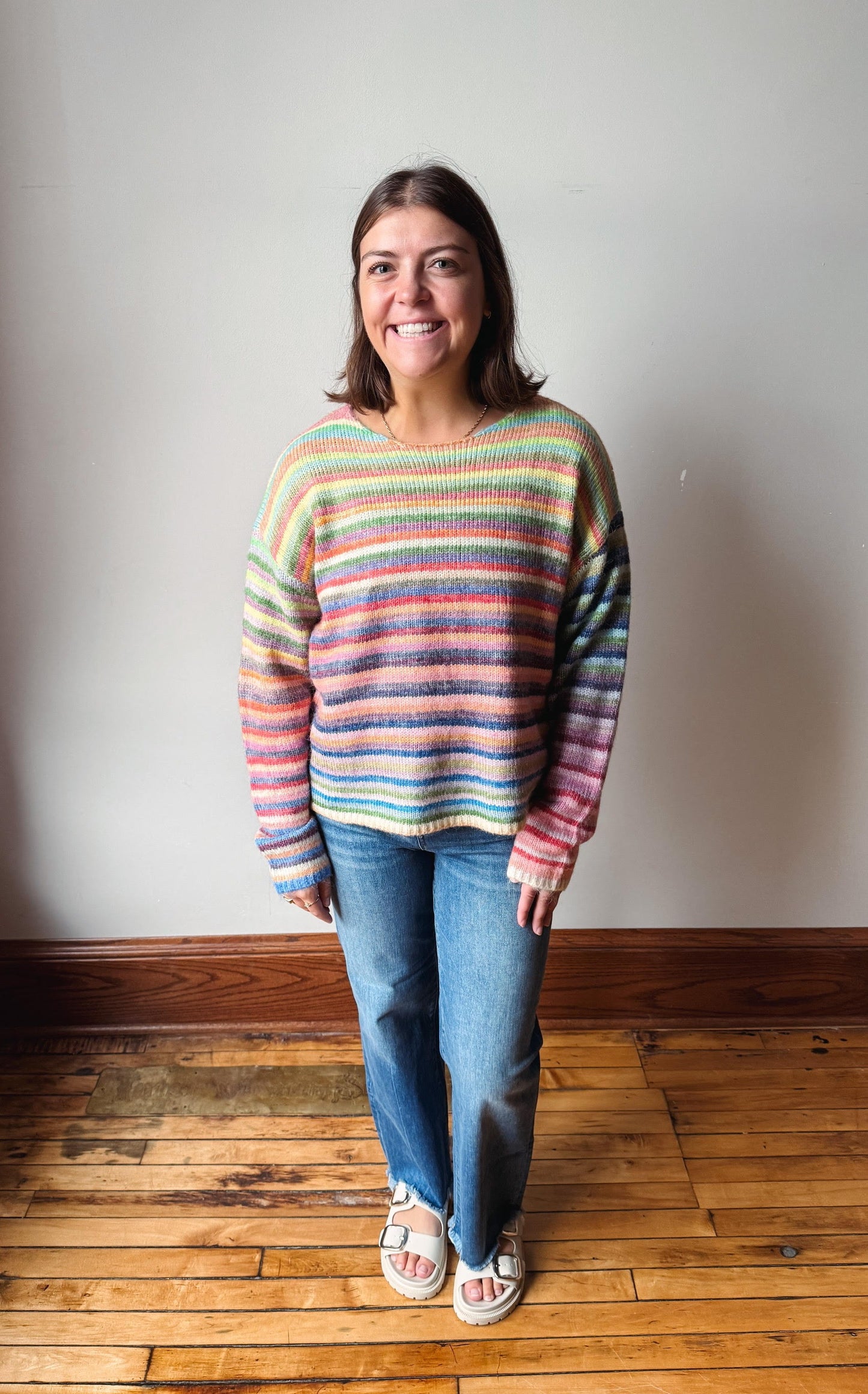 A woman stands in a room with a light gray wall and wooden floor, wearing a colorful horizontally striped knit sweater, blue jeans, and white slide sandals.