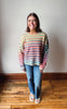 A woman stands in a room with a light gray wall and wooden floor, wearing a colorful horizontally striped knit sweater, blue jeans, and white slide sandals.