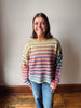 A smiling woman with shoulder-length brown hair stands indoors wearing a colorful horizontally striped knit sweater and blue jeans against a plain light wall and wood floor.
