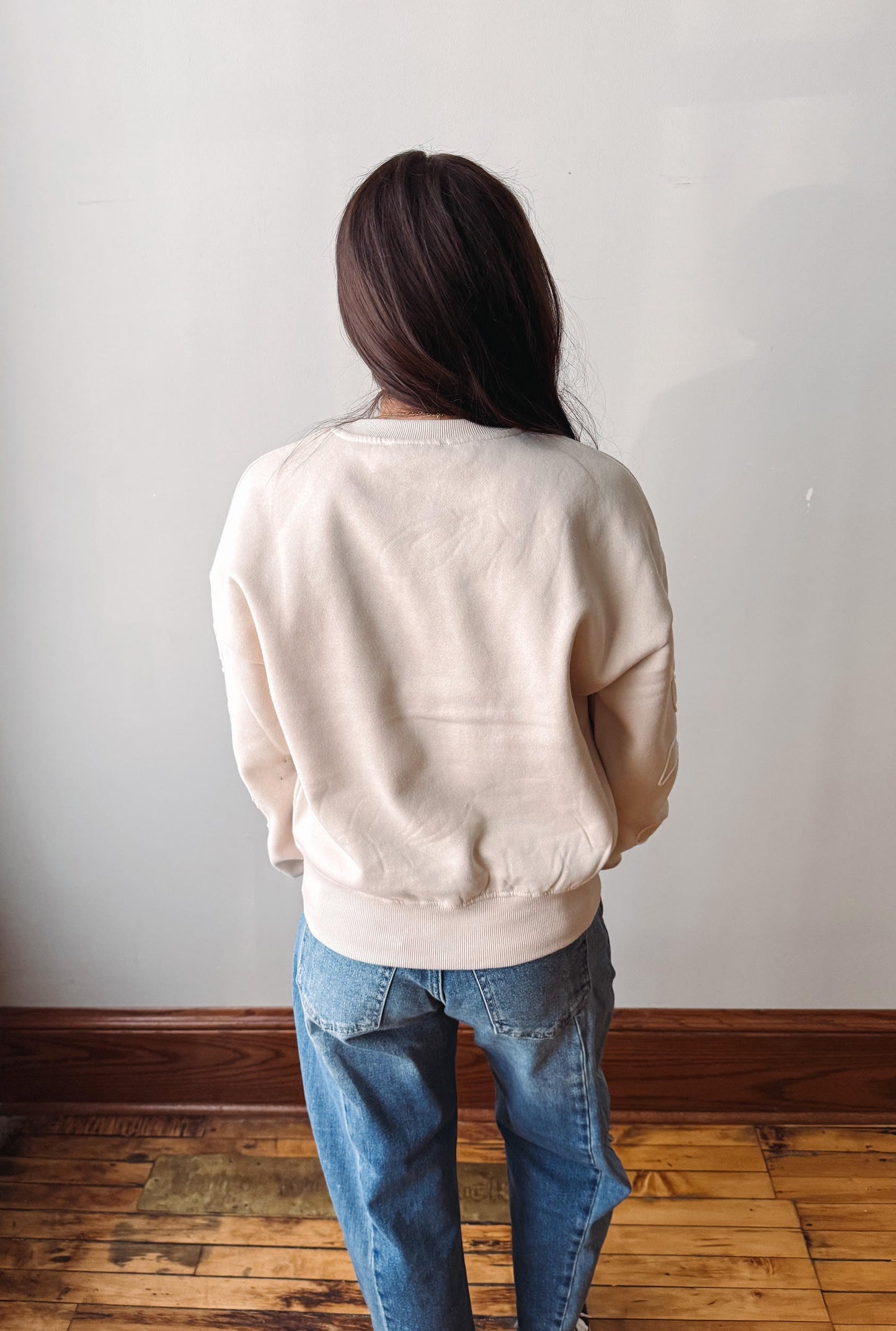 A person viewed from behind wearing a cream-colored crewneck sweatshirt and blue jeans, standing on a wooden floor against a plain light gray wall.