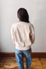 A person viewed from behind wearing a cream-colored crewneck sweatshirt and blue jeans, standing on a wooden floor against a plain light gray wall.