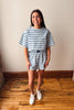 A woman modeling a matching two-piece loungewear set consisting of a short-sleeve crewneck t-shirt and shorts, both featuring a light blue and white horizontal stripe pattern. She is standing on a wooden floor against a plain beige wall.