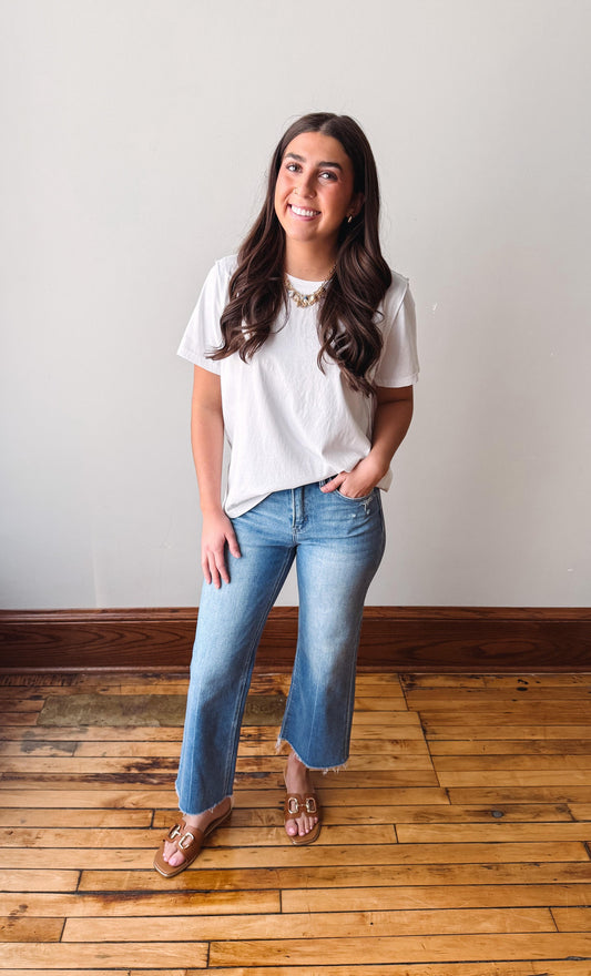 Woman wearing a white t-shirt and blue jeans standing on a wooden floor with a white wall background