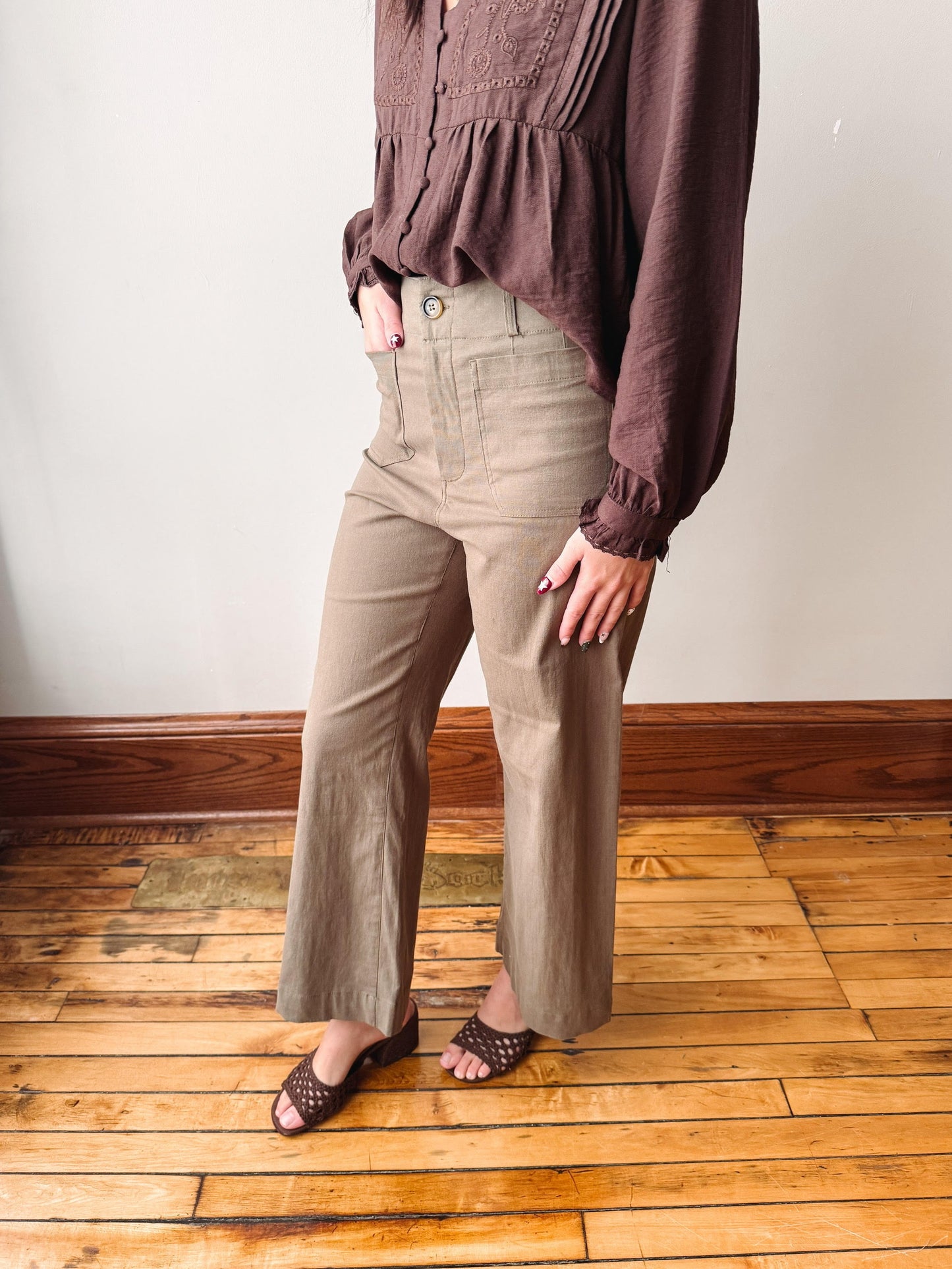 A person wearing high-waisted, wide-leg taupe pants with a matching brown long-sleeve blouse and brown woven sandals, standing on a wooden floor against a white wall.