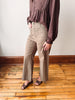 A person wearing high-waisted, wide-leg taupe pants with a matching brown long-sleeve blouse and brown woven sandals, standing on a wooden floor against a white wall.