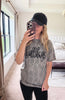 A woman takes a mirror selfie in a bedroom while wearing a gray acid-wash t-shirt with 'bulldogs' printed in black script, a black baseball cap, and black pants.