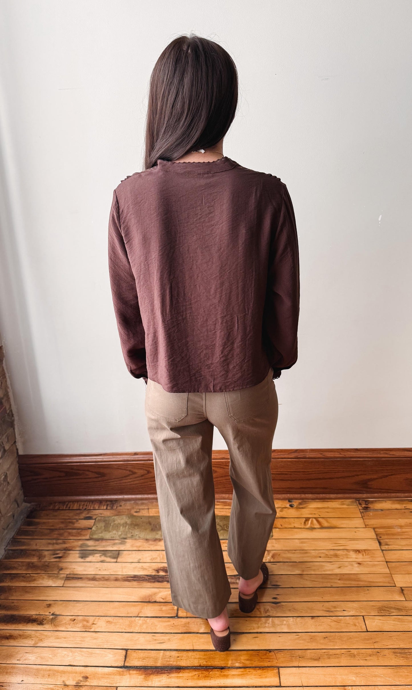 A woman viewed from behind wearing a dark brown long-sleeve top and beige wide-leg pants, standing on a wooden floor against a plain white wall.