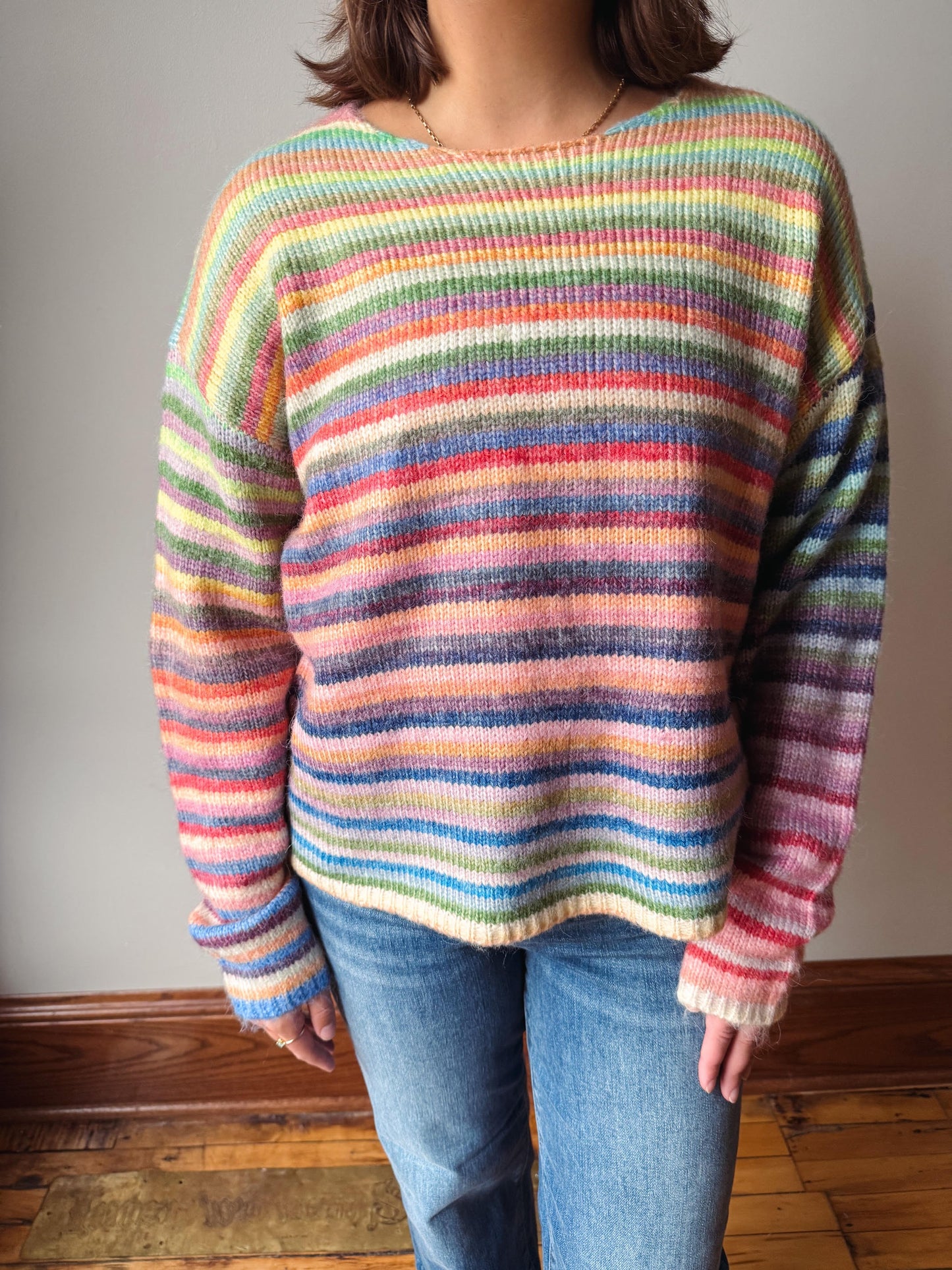 A person wearing a long-sleeve knit sweater with horizontal pastel rainbow stripes in blue, pink, orange, yellow, green, and purple, paired with blue jeans, standing indoors against a plain wall.