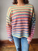 A person wearing a long-sleeve knit sweater with horizontal pastel rainbow stripes in blue, pink, orange, yellow, green, and purple, paired with blue jeans, standing indoors against a plain wall.