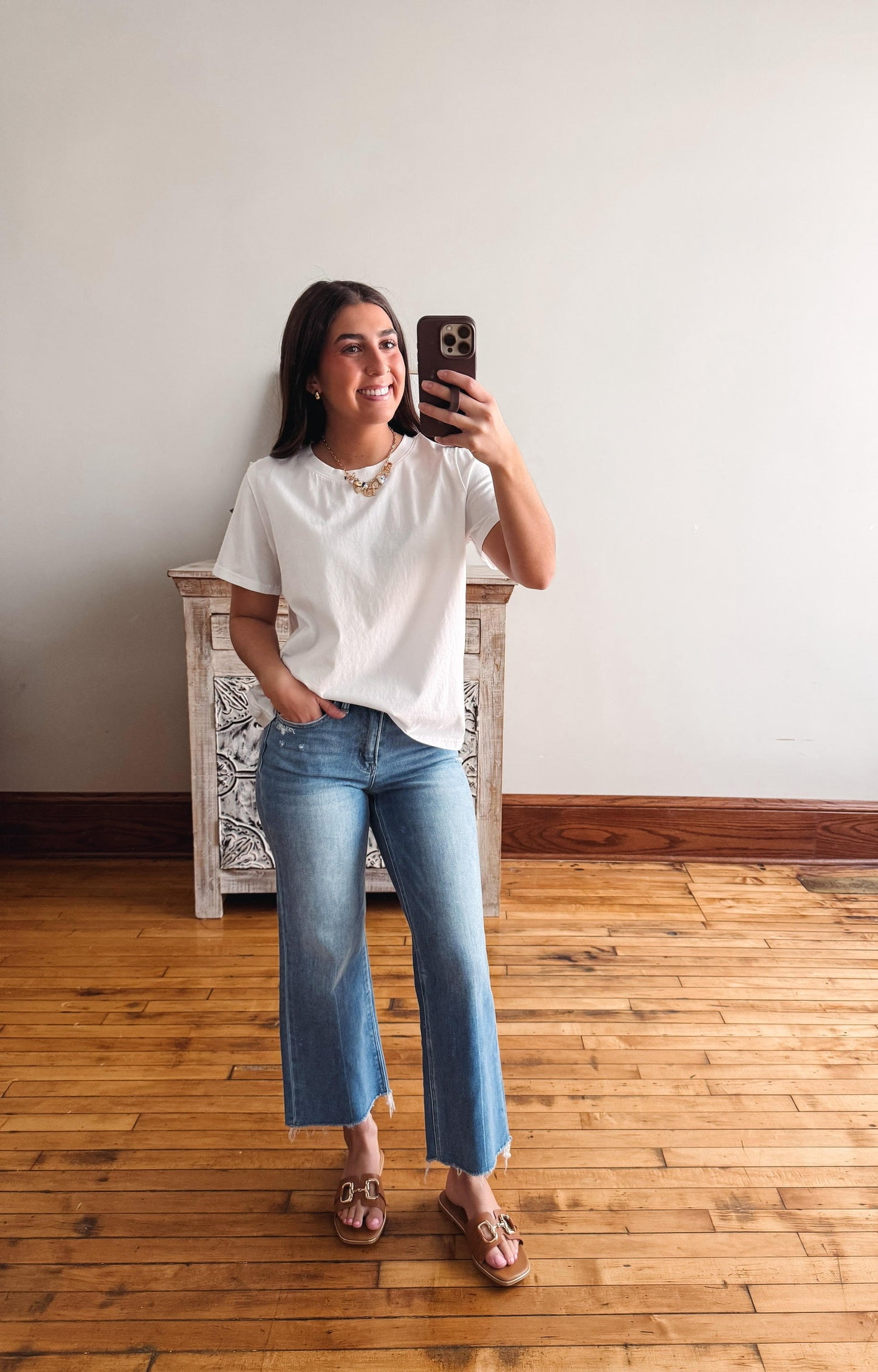 Woman taking a mirror selfie wearing a white blouse and blue jeans in a room with wooden flooring.