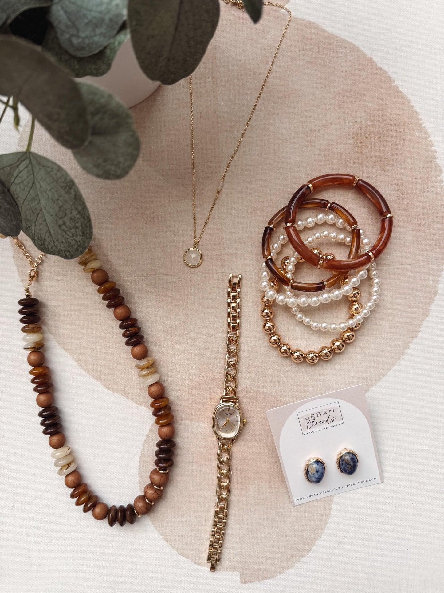 A styled flat-lay of jewelry and accessories on a light beige fabric background with a pink circular accent. Visible items include a beaded necklace, a gold-tone watch, a stack of pearl and brown bangles, a delicate necklace, and a pair of blue-and-gold stud earrings on a branded display card.