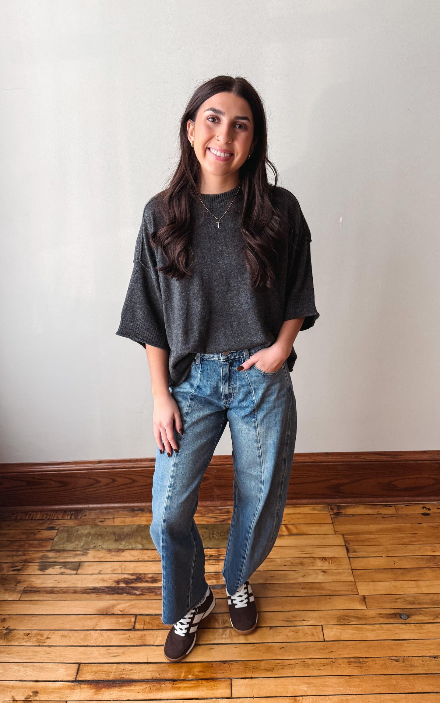 A woman stands in a room with a white wall and wooden floor, wearing a dark gray oversized t-shirt, blue jeans, and brown sneakers.