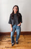 A woman stands in a room with a white wall and wooden floor, wearing a dark gray oversized t-shirt, blue jeans, and brown sneakers.
